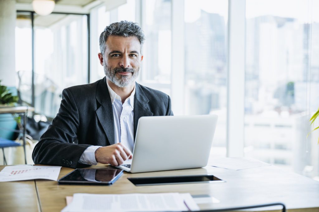 Buenos Aires businessman sitting at office work table next to window with view of downtown and pausing from typing on laptop to smile at camera.