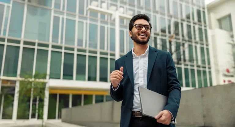 Smiling businessman in suit with laptop near modern office building is looking at side