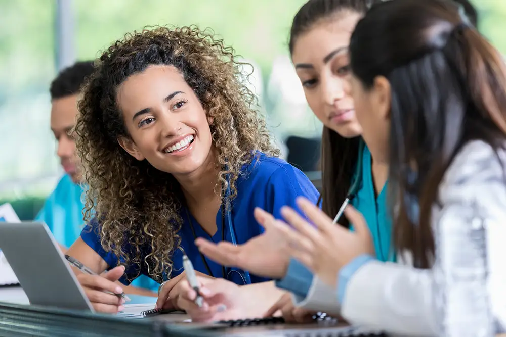 Young woman in class attentively listens, working on medical training assignment with two others.