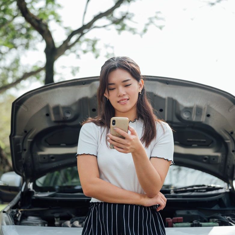 Smiling Young Asian woman waiting car assistant or someone for help with her broken car
while standing near her car with open hood on the road side.